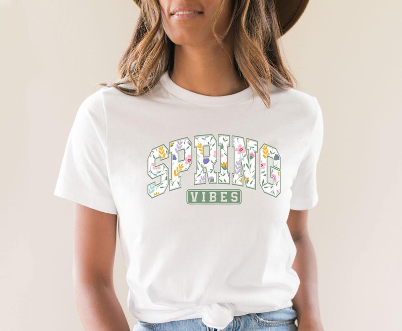 May include: White t-shirt featuring the text "SPRING VIBES" in a floral design. The text is in a collegiate-style font, with each letter filled with colorful flowers and leaves. A green rectangle below the text reads "VIBES".