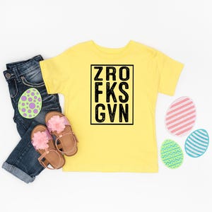 May include: Yellow short-sleeved t-shirt with black text that reads "ZRO FKS GVN". The shirt is laid flat on a white surface with blue jeans, pink sandals, and colorful Easter eggs.