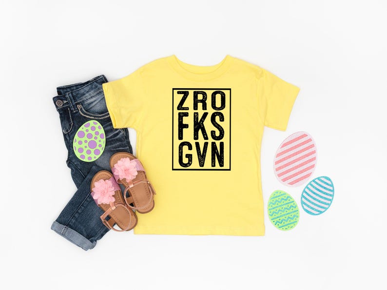 May include: Yellow short-sleeved t-shirt with black text that reads "ZRO FKS GVN". The shirt is laid flat on a white surface with blue jeans, pink sandals, and colorful Easter eggs.