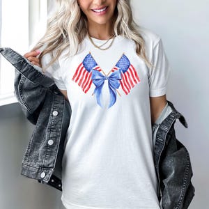 Puede incluir: Camiseta blanca con un dise&ntilde;o de acuarela de dos banderas americanas y un lazo azul. Las banderas tienen rayas rojas y blancas y campos de estrellas azules. El lazo es de color azul claro. La camiseta se usa con una chaqueta vaquera.