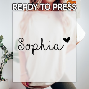 May include: Cream-colored t-shirt with the name "Sophia" in a cursive font and a black heart. The words "READY TO PRESS" are at the top. The shirt is displayed on a person.