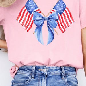 Puede incluir: Camiseta rosa con un dise&ntilde;o de acuarela de dos banderas americanas cruzadas con un lazo azul. Las banderas tienen rayas rojas y blancas y campos de estrellas azules. La camiseta se combina con vaqueros azules.