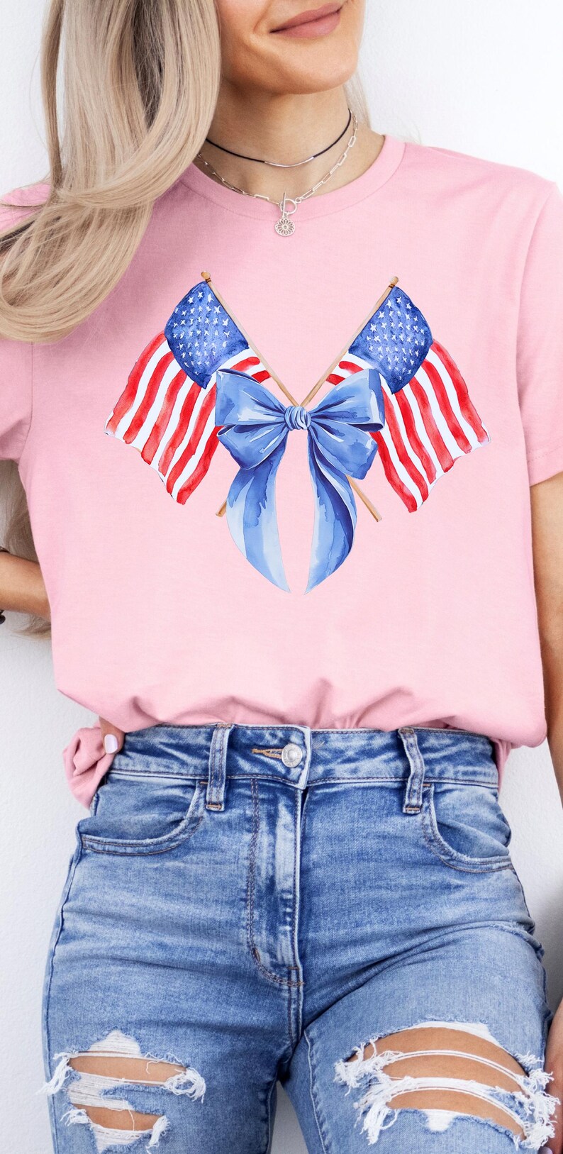 Puede incluir: Camiseta rosa con un dise&ntilde;o de acuarela de dos banderas americanas cruzadas con un lazo azul. Las banderas tienen rayas rojas y blancas y campos de estrellas azules. La camiseta se combina con vaqueros azules.