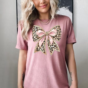 May include: A pink t-shirt with a large leopard print bow with pink ribbon and gold accents.