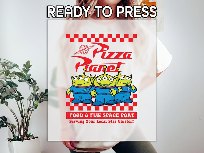 May include: White t-shirt with a red and white checkered design featuring the words "Pizza Planet" in red and three green alien characters from the movie Toy Story. The text "Food & Fun Space Port" and "Serving Your Local Star Cluster!" are also printed on the shirt.