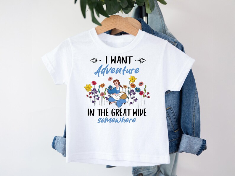 May include: White t-shirt featuring a blue and white illustration of a girl reading a book in a field of flowers. The text on the shirt reads "... I WANT Adventure in the great wide somewhere."