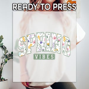 May include: A cream t-shirt featuring the words "SPRING VIBES" in a floral design. The text is in a collegiate style font with a green outline. The words "READY TO PRESS" are at the top.