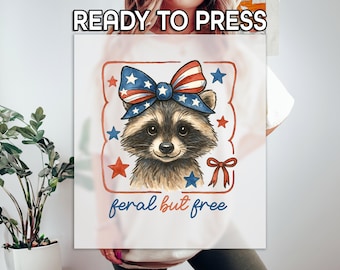 May include: A white t-shirt with a red, white, and blue raccoon wearing a bow with stars and stripes. The raccoon is inside a red frame with stars and the text "feral but free".