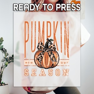 May include: White t-shirt with a fall-themed graphic. The design features the words "PUMPKIN SEASON" in orange, with a pumpkin and a leopard print bow. The words "READY TO PRESS" are at the top.