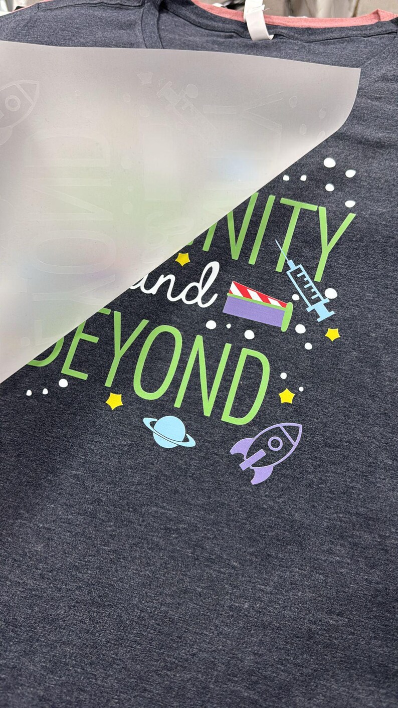 May include: Dark gray t-shirt with the phrase "Infinity and Beyond" in green and white, with space-themed graphics. The design includes a rocket, planet, stars, and a syringe.