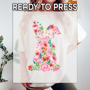 May include: A white t-shirt with a floral design in shades of pink, red, and blue. The design is shaped like a ballerina shoe with a pink ribbon.