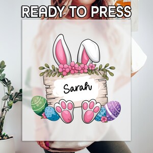 May include: A white graphic featuring bunny ears, pink flowers, and a wooden sign with the name "Sarah". Easter eggs and bunny paws are also included. The text "READY TO PRESS" is at the top.