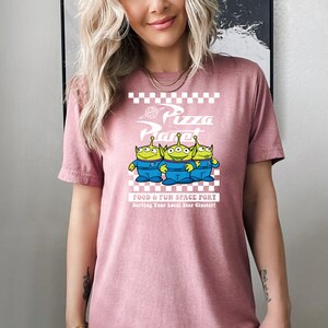 May include: A pink t-shirt with a white graphic of three green aliens from the movie Toy Story. The graphic says "Pizza Planet" with a checkered background and the text "Food & Fun Space Port Serving Your Local Star Cluster!"