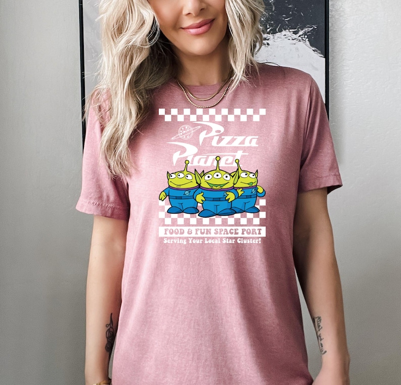 May include: A pink t-shirt with a white graphic of three green aliens from the movie Toy Story. The graphic says "Pizza Planet" with a checkered background and the text "Food & Fun Space Port Serving Your Local Star Cluster!"