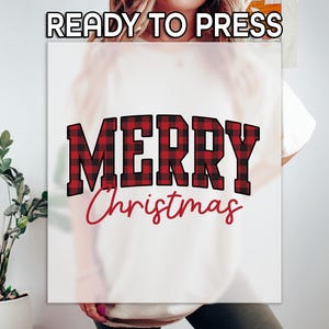 May include: White t-shirt with the words "READY TO PRESS" at the top. The word "MERRY" is in large, block letters with a red and black plaid pattern. The word "Christmas" is written in a cursive font in red.