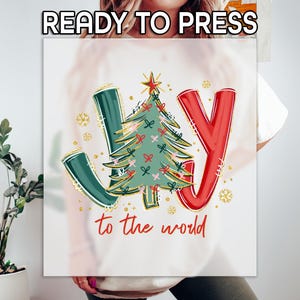 May include: White graphic t-shirt with the words "JOY" in green and red, and a Christmas tree design. The text "to the world" is written in red script. Gold snowflakes and the text "READY TO PRESS" are also visible.