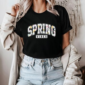 May include: Black t-shirt featuring the text "SPRING VIBES" in a floral design. The word "SPRING" is arched across the top, and "VIBES" is in a rectangular box below. The shirt has a crew neck.