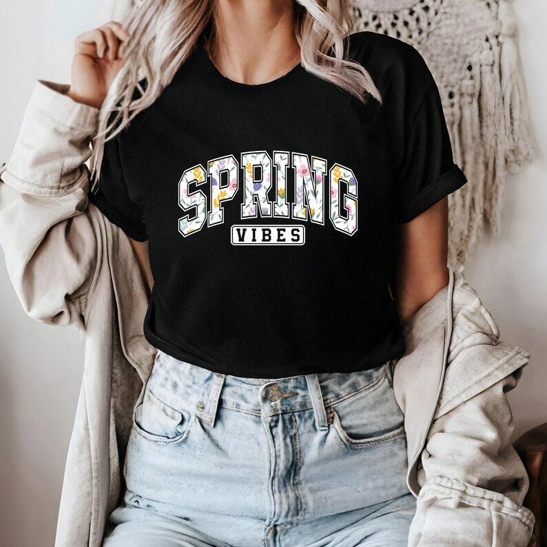 May include: Black t-shirt featuring the text "SPRING VIBES" in a floral design. The word "SPRING" is arched across the top, and "VIBES" is in a rectangular box below. The shirt has a crew neck.