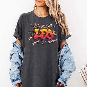 K&ouml;nnte beinhalten: Dunkelgraues T-Shirt mit farbenfrohem Grafikdesign. Das Design zeigt das Wort "LEO" in einem Graffiti-Stil, mit einer Krone dar&uuml;ber und den W&ouml;rtern "Power", "Glamour", "Vitality", "Warm" und "Jovial" drumherum.