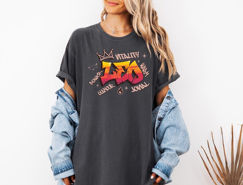 K&ouml;nnte beinhalten: Dunkelgraues T-Shirt mit farbenfrohem Grafikdesign. Das Design zeigt das Wort "LEO" in einem Graffiti-Stil, mit einer Krone dar&uuml;ber und den W&ouml;rtern "Power", "Glamour", "Vitality", "Warm" und "Jovial" drumherum.
