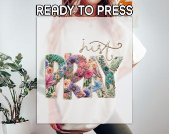Könnte beinhalten: Eine weiße Leinwand mit den Worten "READY TO PRESS" oben. Die Worte "just PRAY" werden in einem floralen Design mit verschiedenen Farben, darunter Rosa, Blau und Grün, angezeigt. Das Wort "just" ist in einer Schreibschrift.