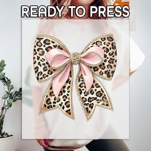 May include: A white t-shirt with a pink and brown leopard print bow with rhinestones. The bow is centered on the shirt. "READY TO PRESS" is written in black text at the top of the image.