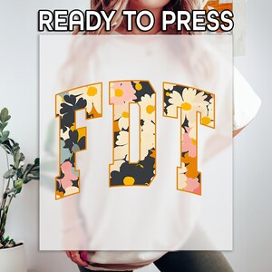 May include: White sweatshirt with the letters "FDT" in a floral design. The letters are outlined in a gold color and filled with a pattern of flowers in shades of pink, yellow, black, and blue. The words "READY TO PRESS" are at the top.