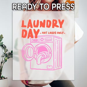 May include: White t-shirt with a pink and orange graphic. The graphic says "LAUNDRY DAY - HOT LOADS ONLY" with a drawing of a washing machine. The words "READY TO PRESS" are at the top of the image.