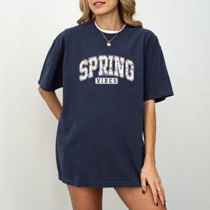 May include: Navy blue t-shirt featuring the text "SPRING VIBES" in a white, floral design. The top has short sleeves and a loose fit. The model is wearing a gold necklace.