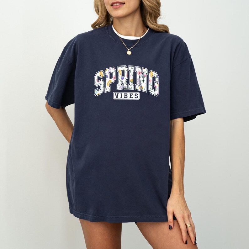 May include: Navy blue t-shirt featuring the text "SPRING VIBES" in a white, floral design. The top has short sleeves and a loose fit. The model is wearing a gold necklace.