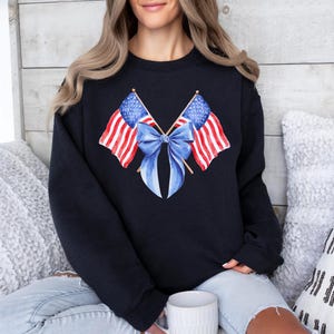 Puede incluir: Sudadera negra con un dise&ntilde;o de acuarela de dos banderas americanas cruzadas con un lazo azul. Las banderas tienen rayas rojas y blancas y campos de estrellas azules. La sudadera es de cuello redondo.