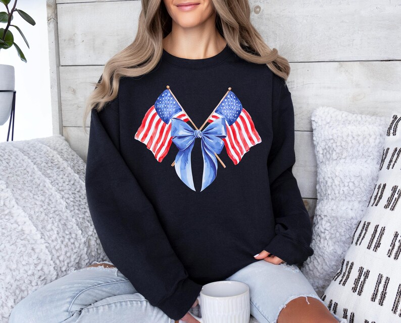 Puede incluir: Sudadera negra con un dise&ntilde;o de acuarela de dos banderas americanas cruzadas con un lazo azul. Las banderas tienen rayas rojas y blancas y campos de estrellas azules. La sudadera es de cuello redondo.