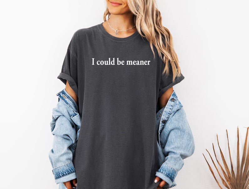 Puede incluir: Una camiseta gris oscuro con el texto "I could be meaner" impreso en blanco en la parte delantera. La camiseta la lleva puesta una persona con el pelo largo rubio y una chaqueta vaquera.