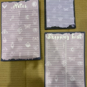 May include: Three grey and black notepad sets with a white, mystical design. The top notepad says "Notes" and the bottom notepad says "Shopping List".