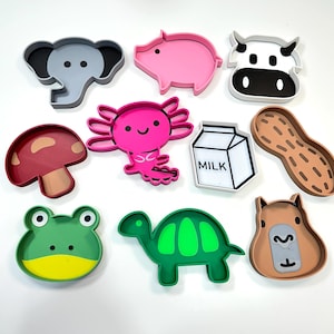 May include: A collection of colorful, cartoon-style animal and food-shaped trays. Includes an elephant, pig, cow, axolotl, mushroom, milk carton, peanut, frog, turtle, and capybara. Each tray has a raised edge.