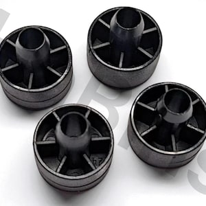 Puede incluir: Cuatro componentes de plástico negro con forma cilíndrica y un agujero central. Cada componente tiene una base circular con soportes radiales. Están dispuestos sobre una superficie blanca. Aproximadamente 2.5 cm de diámetro.