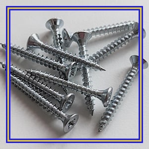 Op de afbeelding: Close-up van een stapel zilverkleurige houtschroeven. De schroeven hebben een kruiskopsleuf en een puntige punt. De schroeven liggen verspreid op een witte ondergrond.