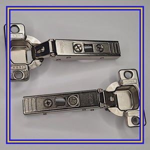 Puede incluir: Dos bisagras plateadas para armarios con la marca "Blum" y el texto "16mm". Cada bisagra tiene un cuerpo rectangular con orificios para tornillos y un brazo giratorio. Diseñadas para puertas de armario.