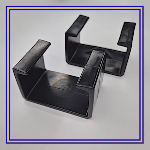 May include: Two black plastic U-shaped brackets. Each bracket has a rectangular base and two upright arms. The brackets are designed to hold or support an object. The image is framed with a blue and yellow border.