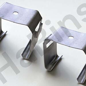 May include: Two silver-colored metal brackets. Each bracket has a rectangular top with a centered hole and curved sides. The brackets are designed with a U-shape and are likely used for construction or mounting purposes.