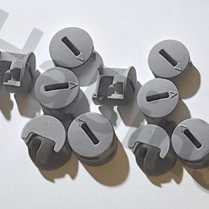 Può includere: Un gruppo di connettori per mobili in plastica grigia. Ogni connettore ha una parte superiore circolare con una fessura e una freccia, e una base curva. Progettati per unire componenti di mobili.