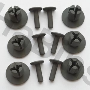 May include: A collection of dark gray plastic fasteners. The fasteners include round-headed rivets and circular pieces with a cross-shaped slot. The pieces are arranged on a white surface. These are likely automotive or industrial components.