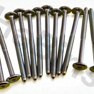 May include: A collection of dark gray metal bolts with flat, circular heads. The bolts have threaded shafts and are arranged on a white surface. The heads have a slightly reflective finish, and the threads are clearly visible.