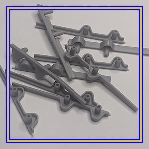 Op de afbeelding: Een verzameling grijze plastic componenten, mogelijk voor een grotere assemblage. De stukken hebben verschillende vormen, waaronder rechte staven en gebogen secties met cirkelvormige uitsteeksels. De onderdelen liggen verspreid over een wit oppervlak.