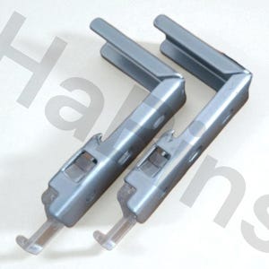 Puede incluir: Dos soportes metálicos de color gris plateado con una pieza de plástico transparente en el extremo. Los soportes tienen forma de L y tienen una serie de agujeros y recortes. El fondo es una superficie blanca lisa.