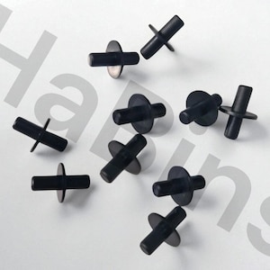 May include: A collection of black plastic fasteners with a cross-shaped head and a circular base, scattered on a white surface. The fasteners are evenly spaced and appear to be new.