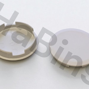 Puede incluir: Dos tapacubos de color gris claro, uno con un diseño abierto y el otro un disco sólido. El tapacubos abierto tiene una base circular con una serie de pestañas y una hendidura central. El tapacubos sólido es un disco liso y redondeado.