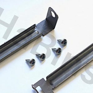Può includere: Primo piano di componenti hardware in metallo, tra cui due staffe rettangolari e quattro viti argentate. Le staffe hanno un design a canale e un piccolo foro per il montaggio. Le viti sono piccole e hanno una testa a forma di stella.