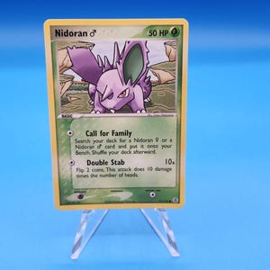 May include: A Nidoran male trading card with 50 HP, featuring a purple creature illustration. The card text includes Call for Family and Double Stab attacks, with a green background border and a yellow frame, displayed on a clear plastic stand.