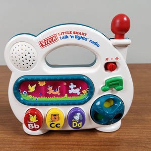 May include: A white VTech Little Smart Talk 'n Lights radio toy with a red antenna, a speaker, and a colorful display screen. It features three round buttons labeled with bird, cat, and dog illustrations, a green musical note button, and a dial.